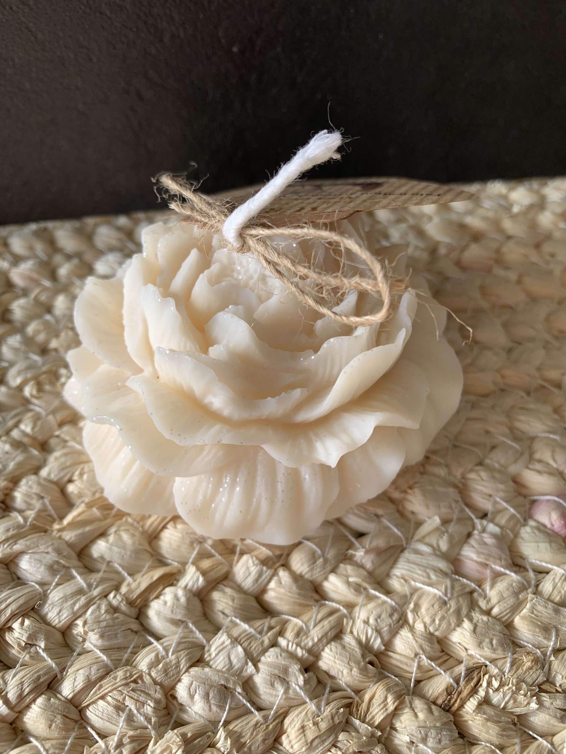 Bougie moulée en forme de pivoine couleur ivoire, finition fleur détaillée, posée sur un tapis tressé naturel avec ficelle et étiquette.